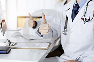 Two doctors sitting with thumbs up in hospital office. Medical help, countering viral infection and medicine concept