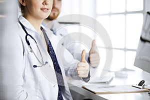 Two doctors sitting with thumbs up in hospital office. Medical help, countering viral infection and medicine concept