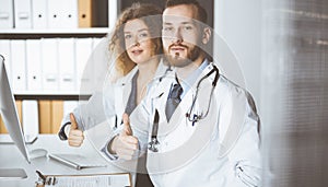 Two doctors sitting with thumbs up in hospital office. Medical help, countering viral infection and medicine concept