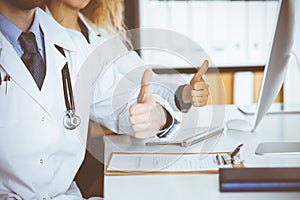 Two doctors sitting with thumbs up in hospital office. Medical help, countering viral infection and medicine concept