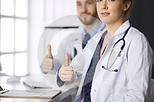 Two doctors sitting with thumbs up in hospital office. Medical help, countering viral infection and medicine concept