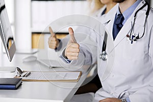 Two doctors sitting with thumbs up in hospital office. Medical help, countering viral infection and medicine concept