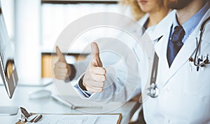 Two doctors sitting with thumbs up in hospital office. Medical help, countering viral infection and medicine concept