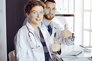 Two doctors sitting with thumbs up in hospital office. Medical help, countering viral infection and medicine concept