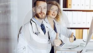 Two doctors sitting with thumbs up in hospital office. Medical help, countering viral infection and medicine concept