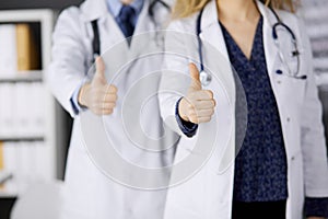 Two doctors standing with thumbs up in hospital office. Medical help, countering viral infection and medicine concept