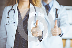 Two doctors standing with thumbs up in hospital office. Medical help, countering viral infection and medicine concept