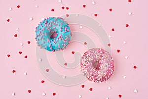 Two donuts decorated icing and sprinkles and confetti in the shape of heart on pink background