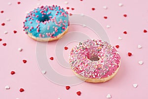 Two donuts decorated icing and sprinkles and confetti in the shape of heart on pink background