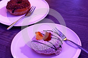 Two donuts in pink with coconut flakes and white chocolate served on a plate and caramel donut in the background on a wooden look