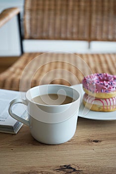 Donuts with pink icing, a cup of coffee with milk and a book on a wooden table against the background of a chair