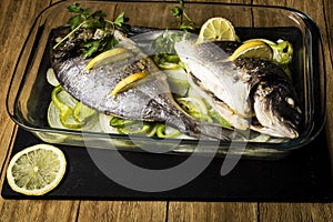 Two Dorada fish cooked in the oven with lemon, onion and green pimento in a glass dish on a black slate tray