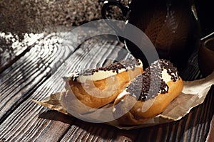 Two eclairs with cream and chocolate chips beside a black mug on a dark wooden surface, softly lit with a rustic backdrop