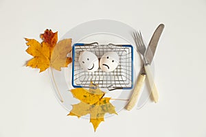 Two eggs with a joyful smile and a sad one lie in a supermarket basket next to autumn leaves and a knife with a fork on