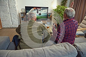 Two elderly men, sport fans sitting on couch at home in living room and watching online football match translation on tv