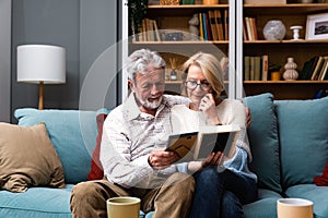 Two elderly people senior couple man and woman looking at a family photo smile and hug while sitting at home on sofa. memories and