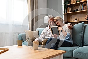 Two elderly people senior couple man and woman looking at a family photo smile and hug while sitting at home on sofa. memories and