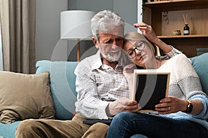 Two elderly people senior couple man and woman looking at a family photo smile and hug while sitting at home on sofa. memories and