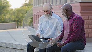 Two Elderly people sitting outdoors using laptop for work. Mature adult African American male counselor helps old