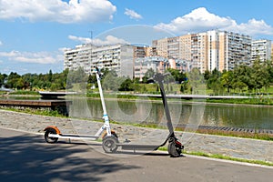 Two electric scooters, white and black, stand on the bandwagon on the street. Sunny summer day. Modern city transport.