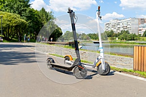 Two electric scooters, white and black, stand on the bandwagon on the street. Sunny summer day. Modern city transport. City park n