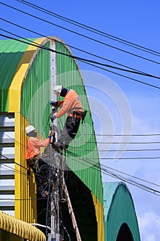 Two electricians installing electric cable lines on power pole near colorful warehouse building against blue sky background