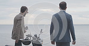 Two elegant Caucasian men walking to river bank in slowmo with scooter steering wheel at front. Back view middle shot of