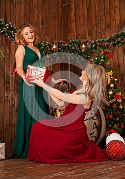 Two elegant women in red and green long evening dress posing near christmas tree indoors