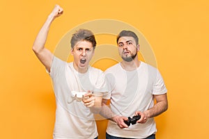 Two emotional friend with joysticks in their hands play video games, isolated on a yellow background. Winner and loser are playing