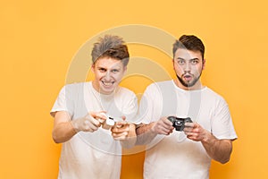 Two emotional gamer with gamepad in hand, playing video games focused on a yellow background looking into the camera. Two friends