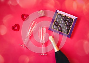Two empty glasses, bottle of champagne, box of chocolate, gift and two red hearts on pink background