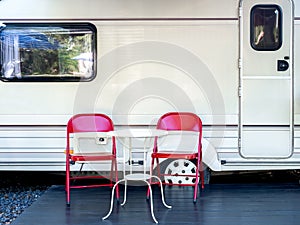 Two empty red iron chairs with white round table in front of the door and window of caravan car with nobody.