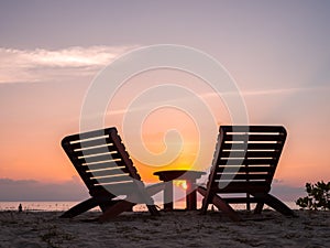 Two empty silhouette wooden sunbeds, couple chair seat and side table on sand beach, peaceful evening sunset sky background with