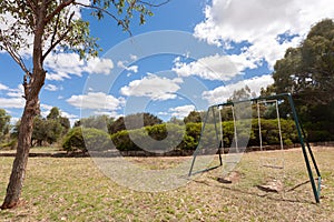 Two empty swings on grass with a small tree in the foreground under a blue sky with some white clouds