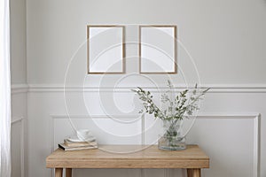 Two empty vertical picture frame mockups hanging on wall. Cup of coffee, books. Wooden desk, table. Vase, green grasses