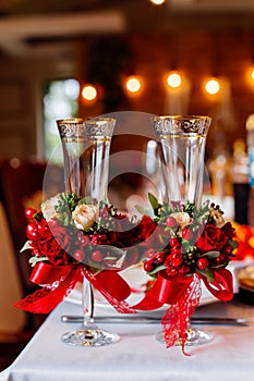 Two empty wedding glasses, decorated with greenery, red roses and ribbon, standing on the banquet table.