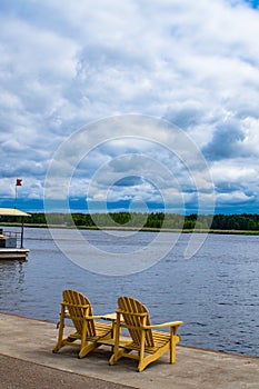 Two empty wooden Adirondack chairs or Muskoka deckchairs on wood shore overlooking scenic calm river