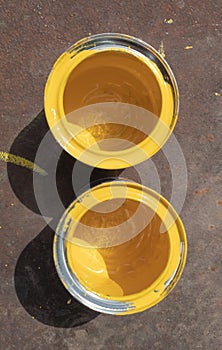 Two empty yellow paint cans with smudges stand on a rusty iron sheet lit by sunlight