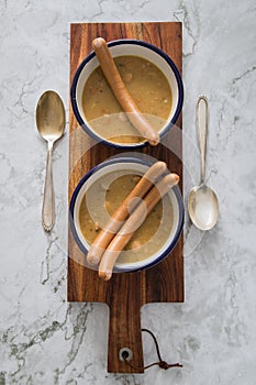 Two enamel bowls hearty German yellow split pea soup stew with wiener sausages on wooden board and light marble background