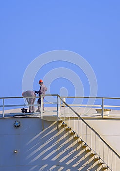 Two engineers checking oil quality on top of oil fuel storage tank against blue sky in vertical frame