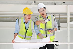 Two engineers man standing in the construction plant. Right engineer holding paper roll and point to construction area