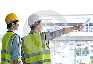 Two engineers man standing in front of the site. Right engineer holding paper roll and point to construction area