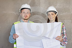 two engineers, a man and a woman in white helmets and protective vests, are standing in the room and holding a plan of