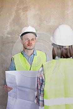 two engineers, a man and a woman in white helmets and protective vests, are standing in the room and holding a plan of