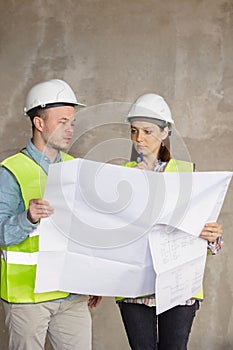 two engineers, a man and a woman in white helmets and protective vests, are standing in the room and holding a plan of