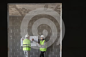 two engineers, a man and a woman in white helmets and protective vests, are standing in the room and holding a plan of