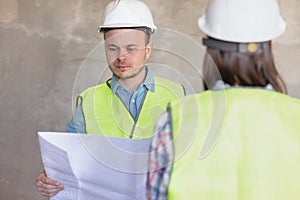 two engineers, a man and a woman in white helmets and protective vests, are standing in the room and holding a plan of