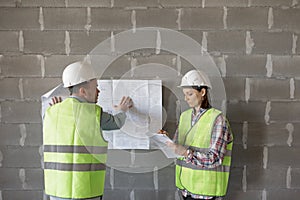 two engineers, a man and a woman in white helmets and protective vests, are standing in the room and holding a plan of