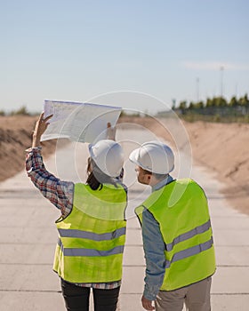 two engineers, a man and a woman in white helmets and protective vests, are standing on the street and holding a plan