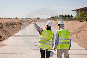 two engineers, a man and a woman in white helmets and protective vests, are standing on the street and holding a plan